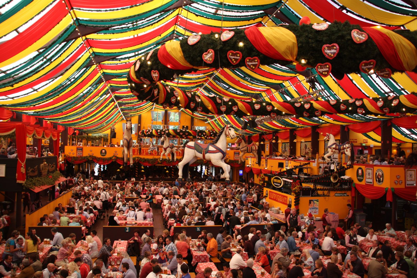 뮌헨 옥토버페스트 히포드롬 천막 안, 빨강·노랑·초록 줄무늬 천장 아래 회전목마 장식과 긴 테이블을 가득 메운 관객