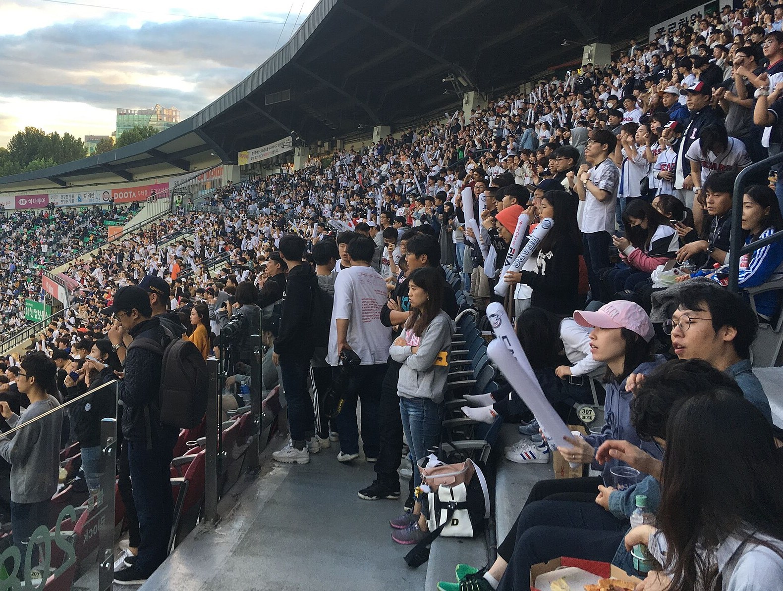 해 질 무렵 서울 잠실야구장 관중석. 흰색·남색 응원 유니폼을 입은 관중이 내야 스탠드를 빽빽하게 메웠고, 앞쪽 통로에 선 사람들이 흰 막대풍선을 들고 경기장을 내려다보고 있다. 왼쪽 위로 구름 낀 저녁 하늘과 건너편 관중석이 보인다